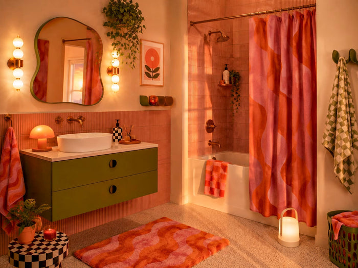 Full bathroom view with olive vanity, curved mirror, pink and orange shower curtain, wavy bath mat and warm lighting.